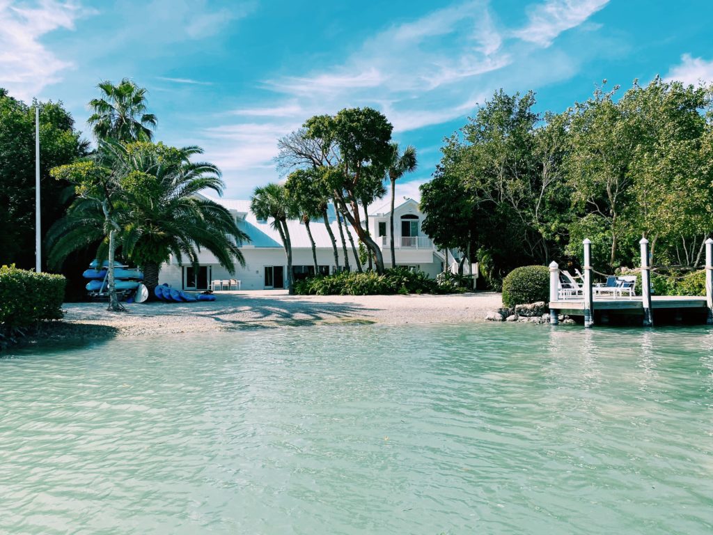 A view of the Sea Oats Estate in Captiva Island, FL from the water near the beach with a dock and kayaks near palm trees