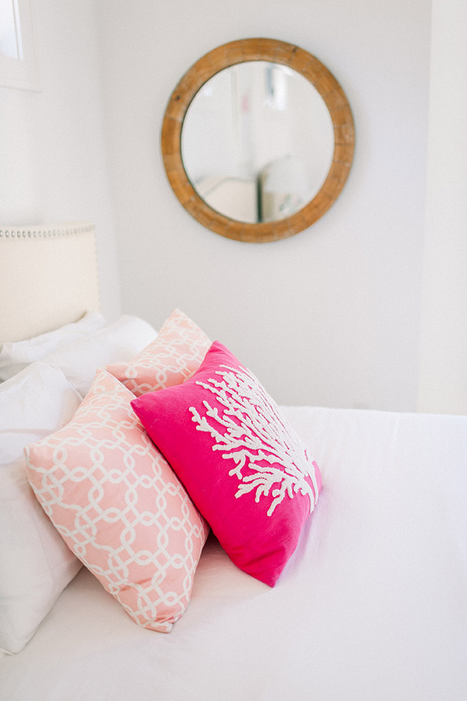 Guest room in the Sea Oats Estate with a queen bed made with white linen sheets two light pink pillows with a white pattern and a hot pink pillow with a texture coral design and a circular wooden framed mirror hung on the wall in Captiva Island, FL