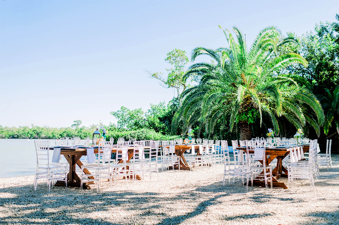 An assortment of brown tables and white chairs on beach with plates, glasses and napkins on top next to ocean and palm trees at the Sea Oats Estate in Captiva Island, FL