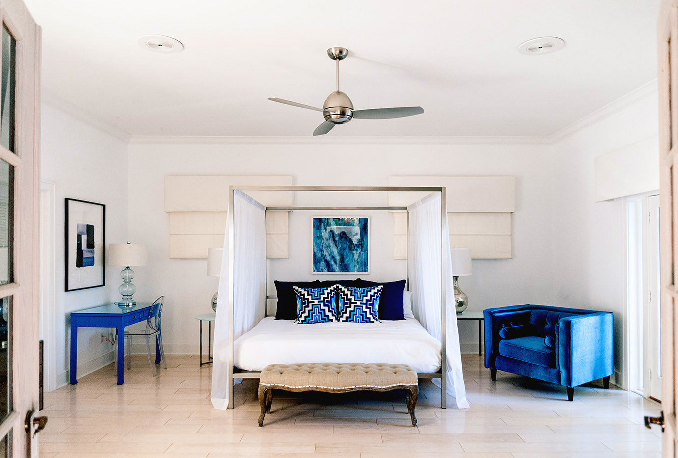 Luxurious guest room with a canopy bed draped with white sheer curtains, white bedding, navy blue and bright blue aztec pillows, a sitting bench at the foot of the bed and a velvet blue lunge chair at the estate in Captiva Island, FL