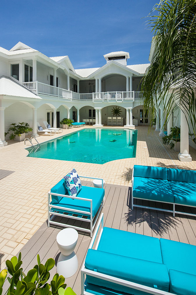 White Estate in Captiva Island, FL with an in-ground pool, blue patio furniture, white pool chairs, a patio area with green plants and a second floor balcony overlooking the pool surrounded by palm trees