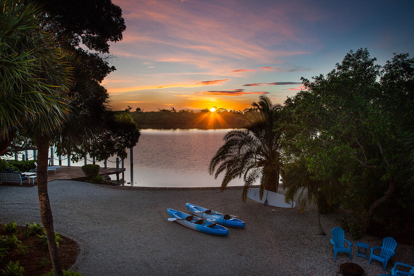 Sunset view from the private Sea Oats Estate with kayaks on the beach and a view of the private dock and a fire pit with blue adirondack chairs