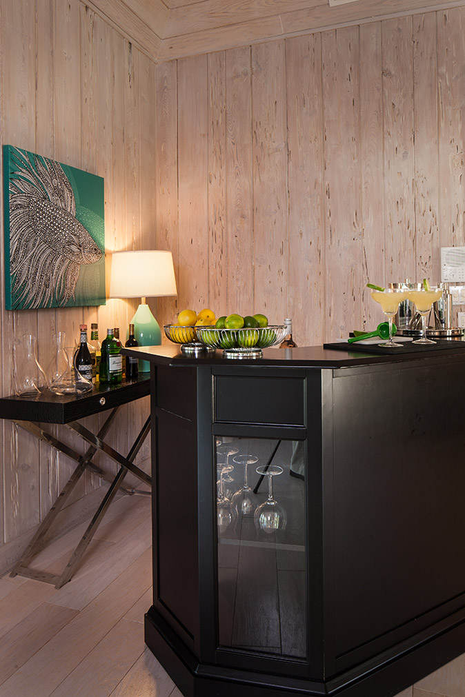 Black wooden bar with a basket of lemons and limes and margaritas on the bar top; a back bar table with wine and liquor on it with decanters and a blue lamp surround by wooden plank walls at Sea Oats Estate in Captiva Island, FL