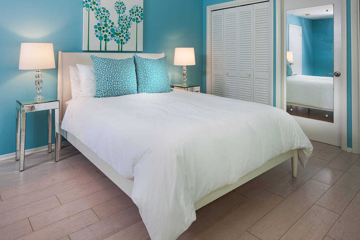 Guest room in the Sea Oats Estate with bright blue walls, a white bed frame, white bedding and pillows, two bright blue decorative pillows, glass nightstands with lamps and a closet with a mirror