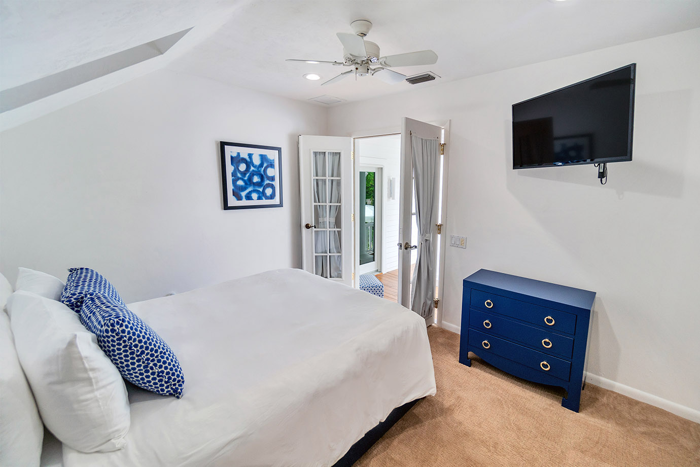 Bedroom in the Captiva Island, FL Sea Oats Estate with a bed with white bedding and pillows, two decorative navy and white pillows, a blue dresser with gold handles, a tv on the wall and french doors leading to the patio