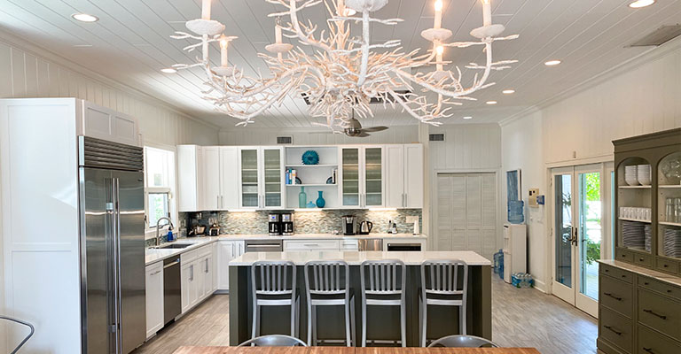 Full equipped kitchen in the Sea Oats Estate in Captiva Island, FL with white cabinetry, stainless steel appliances, a large kitchen island white silver metal barstools and a white coral chandelier