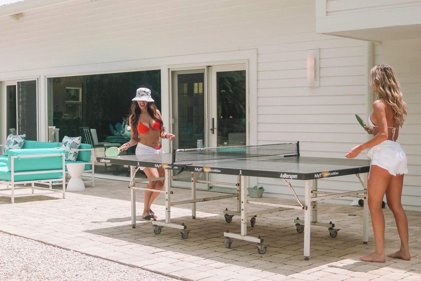 Two women in bathing suits and white shorts playing ping pong outdoors near teal patio furniture at the Sea Oats Estate in Captiva Island, FL