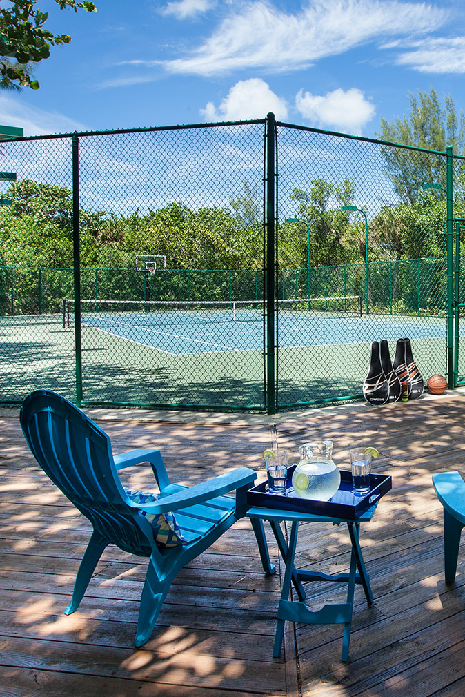 Two bright blue adirondack chairs with a side table of water and lemon in a pitcher; overlooking the fenced off tennis and basketball court at the Sea Oats Estate