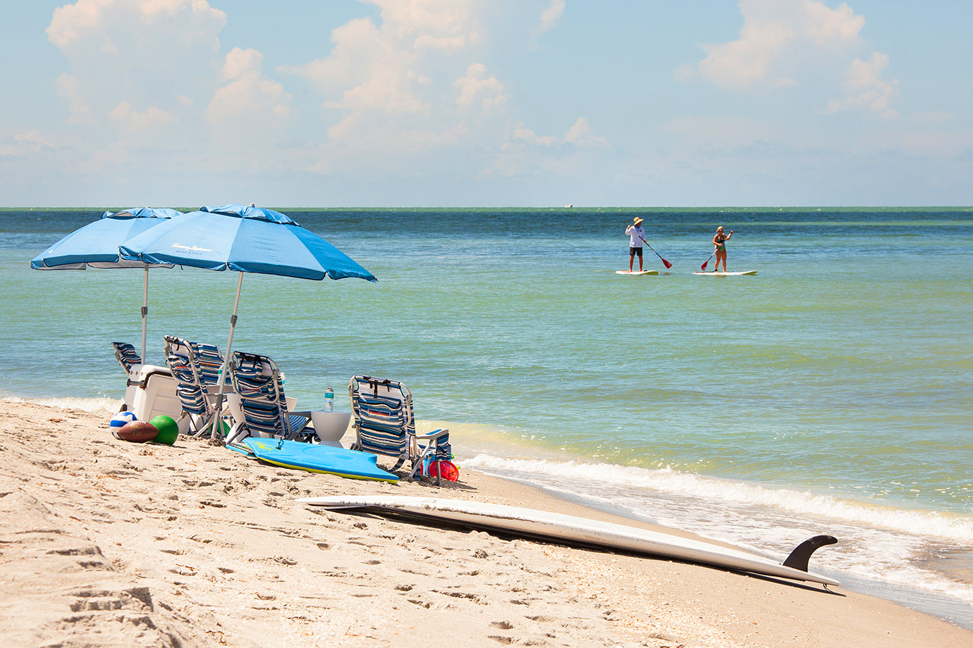 Shore line in Captiva Island, FL with five beach chairs and two umbrellas white two people paddle board in the ocean in the distance
