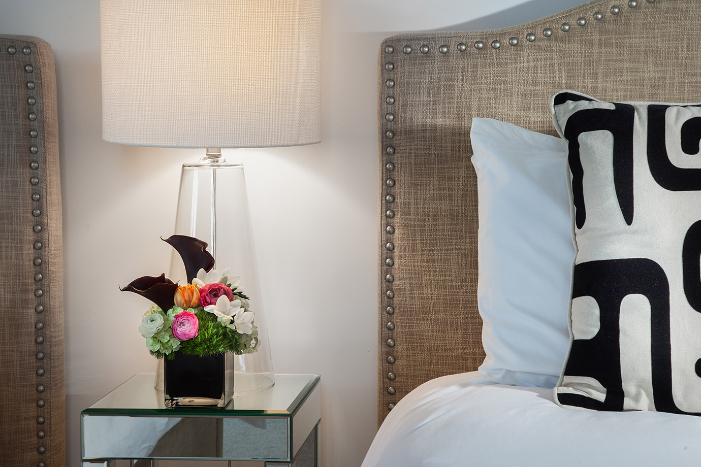 Lamp on Bedside Table behind flowers next to White pillow and a decorative pillow with a black and white pattern in front of a headboard on a bed with white linens at the Sea Oats Estate in Captiva Island, FL