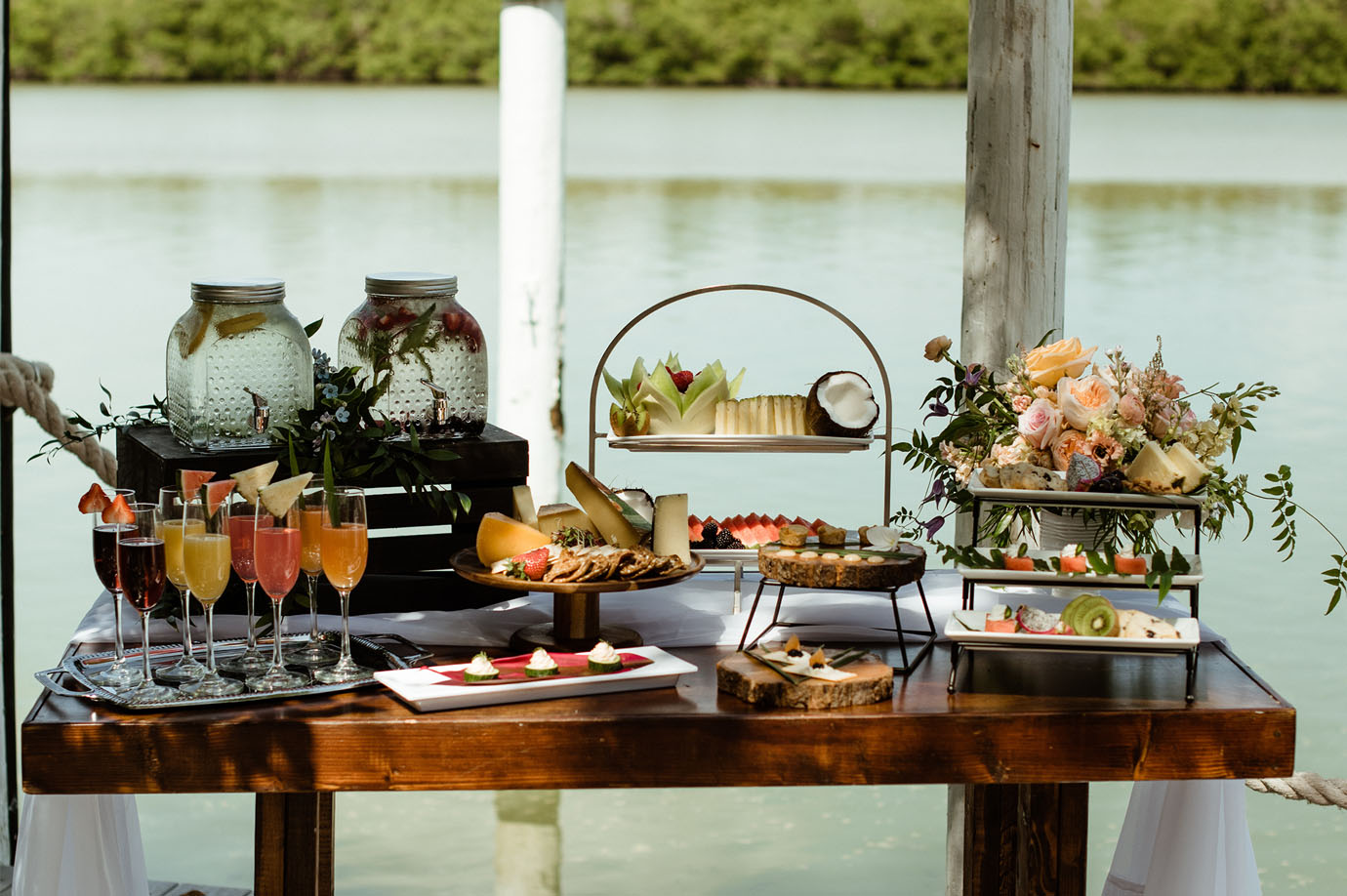 An array of beverages, vegetables and snacks on top of a table with a floral piece in front of the water at the Sea Oats Estate in Captiva Island, FL