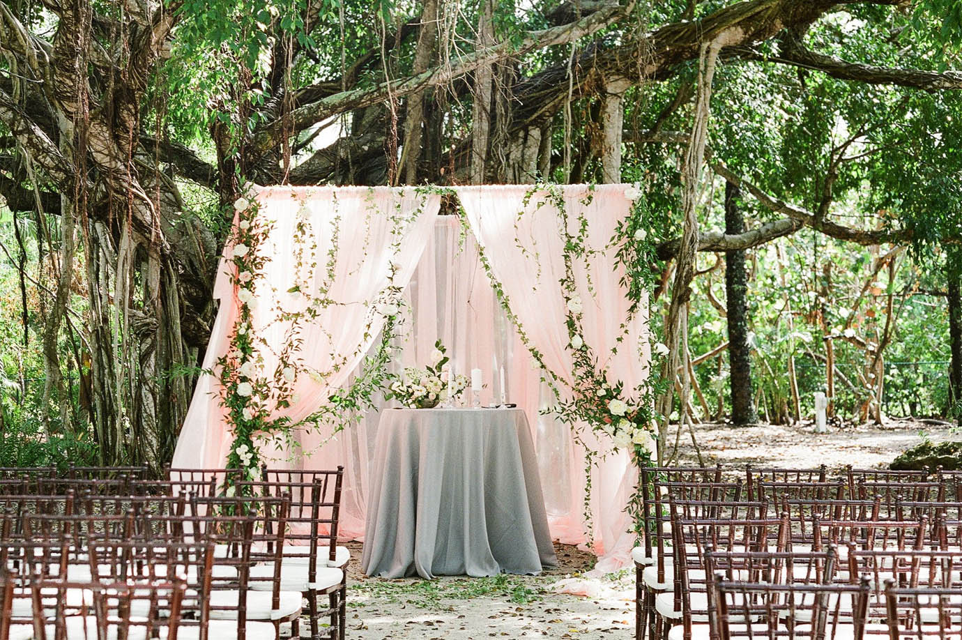 Wedding ceremony set up under a large tree with brown ceremony chairs and a pink backdrop with a grey linen table and greenery strung throughout in Captiva Island, FL