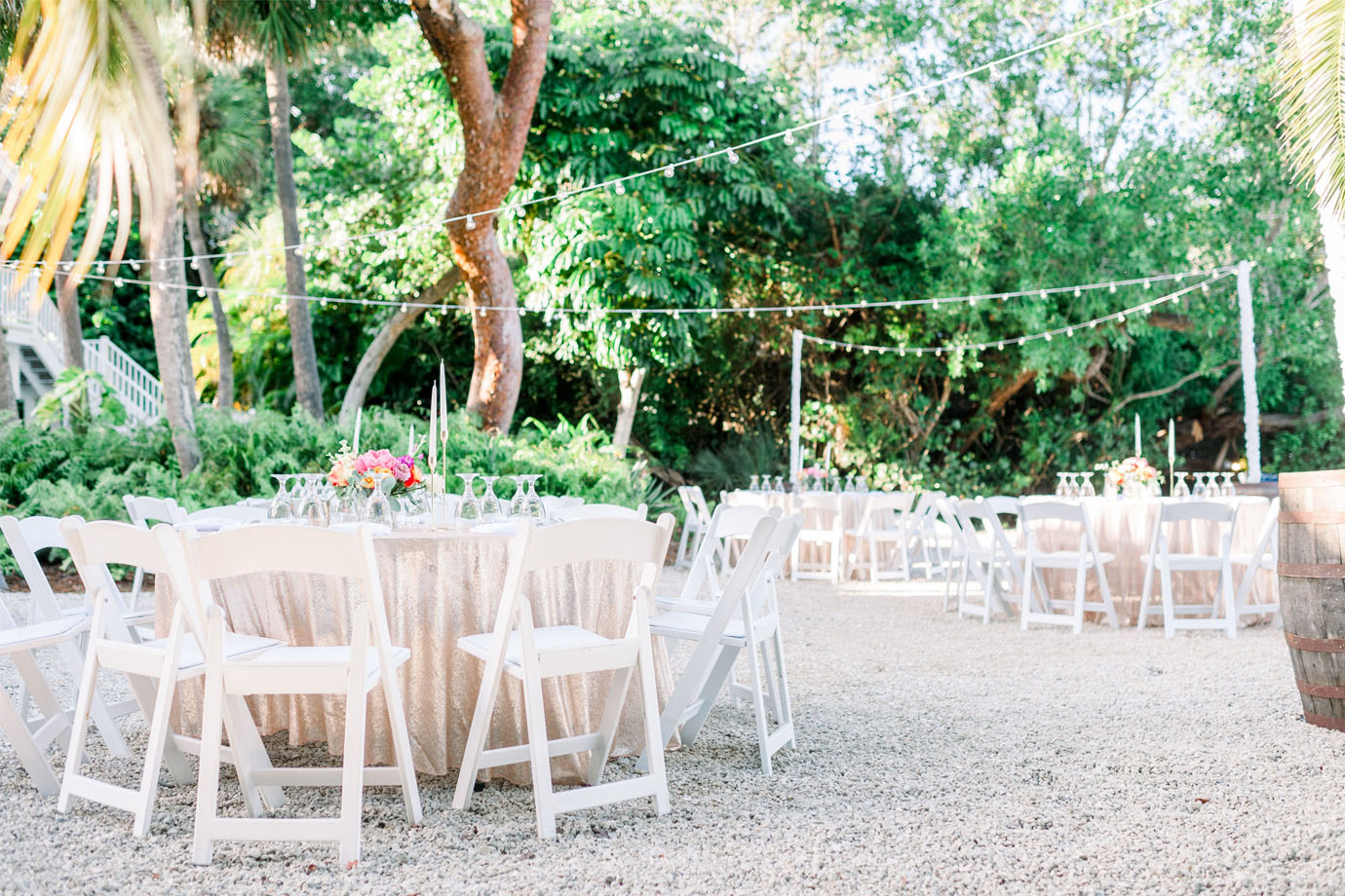 Round guest tables with white folding chairs set white champagne sequin linens and bright pink and orange floral with tall candlesticks; surrounded by palm trees and greenery with bistro lighting strung throughout the reception area in Captiva Island, FL at the Sea Oats Estate