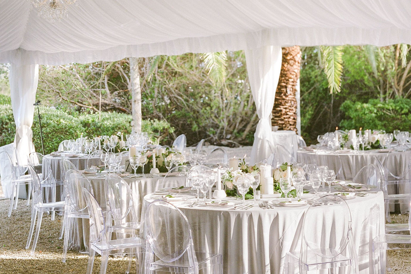 Reception underneath a white cloth tent with round tables set with tan linens and clear chairs with the tables set for dinner service and white floral and greenery at Sea Oats Estate