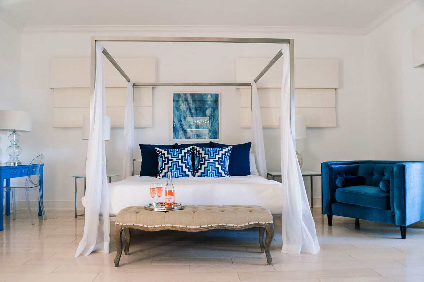 Luxurious guest room with a canopy bed draped with white sheer curtains, white bedding, navy blue and bright blue aztec pillows, a sitting bench at the foot of the bed and a velvet blue lunge chair at the estate in Captiva Island, FL