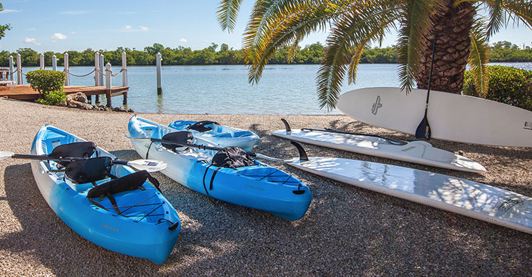 Two blue kayaks and white paddle boards on the shore of the beach next to a wooden dock in Captiva Island, FL