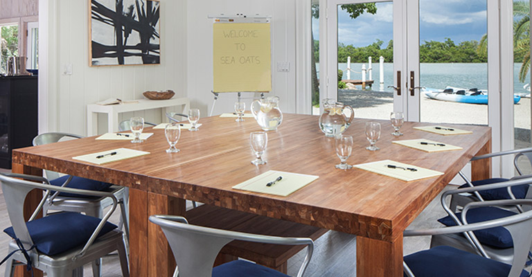 Wooden dining room table with silver metal chairs and navy blue cushions set for a corporate retreat with notepads, pens and water glasses with a view of the ocean at the Sea Oats Estate in Captiva Island, FL