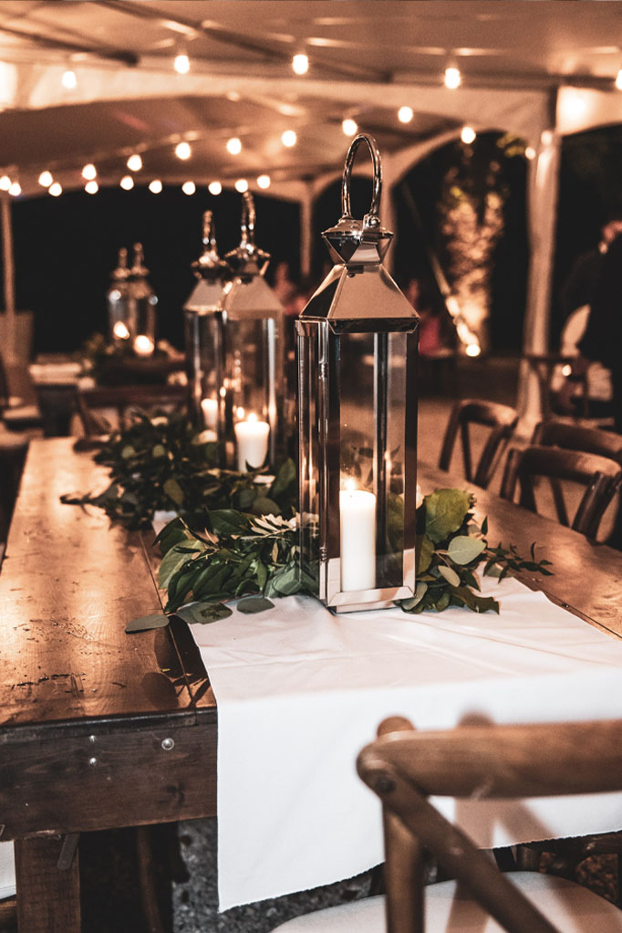 Wedding reception set up under a white tent with brown farm tables and wooden chairs with white runners, tall silver lanterns and greenery down the center of the table in Captiva Island, FL