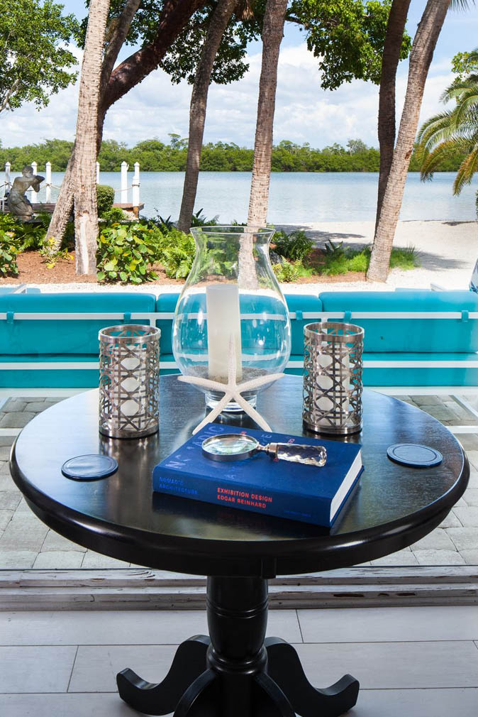 A table with candles, a book, a starfish, coasters and a vase near teal patio furniture in front of palm trees at the Sea Oats Estate in Captiva Island, FL