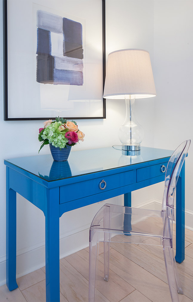 Clear chair in front of blue table with glass top with lamp and flowers on top in front of modern art painting at the Sea Oats Estate in Captiva Island, FL