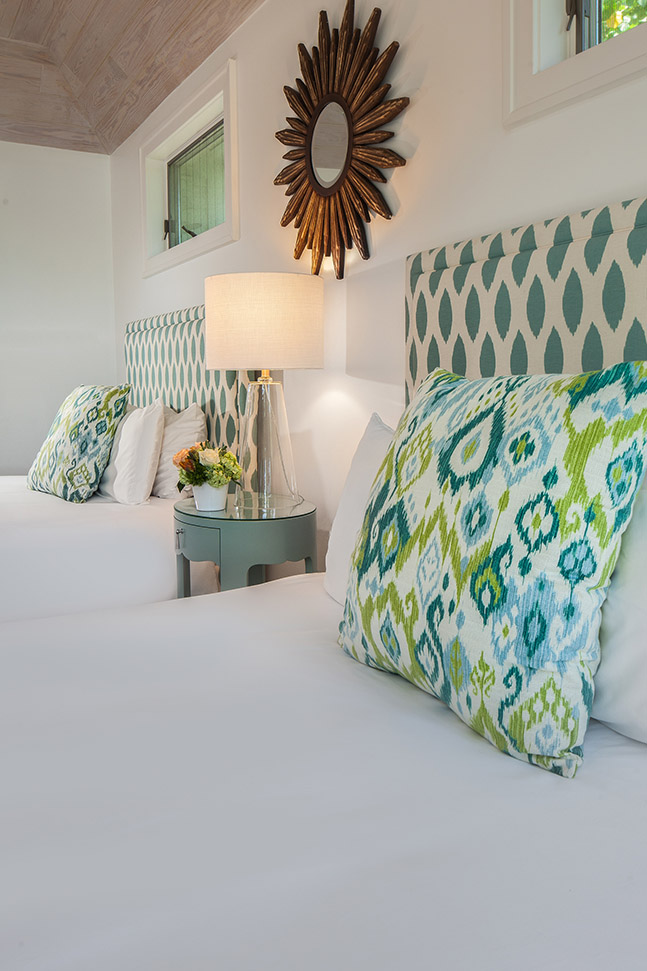 Bedroom in Sea Oats Estate with two queen beds with white linen sheets, blue and white fabric headboard with blue and green throw pillows, a blue nightstand between the beds with a lamp