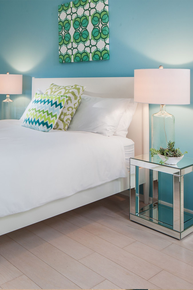 Bedroom in the Sea Oats Estate with white linen sheets and pillows, blue an green textured pillows, a mirror nightstand with a glass lamp and succulent plant and blue walls with a green and blue piece of art on the wall