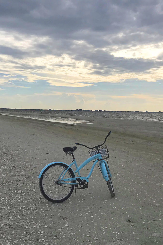 Blue bike with basket on sandy beach near water at low tide with grey clouds and sun above at the Sea Oats Estate in Captiva Island, FL