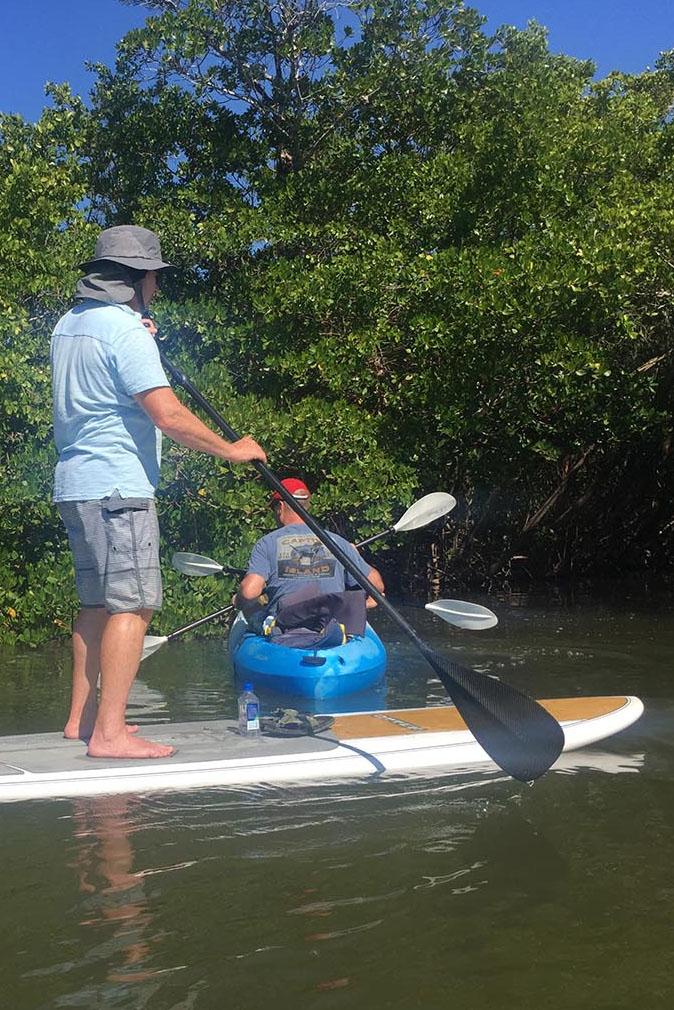 Two men on a paddleboard and kayak surrounded by green trees in Captiva Island, FL