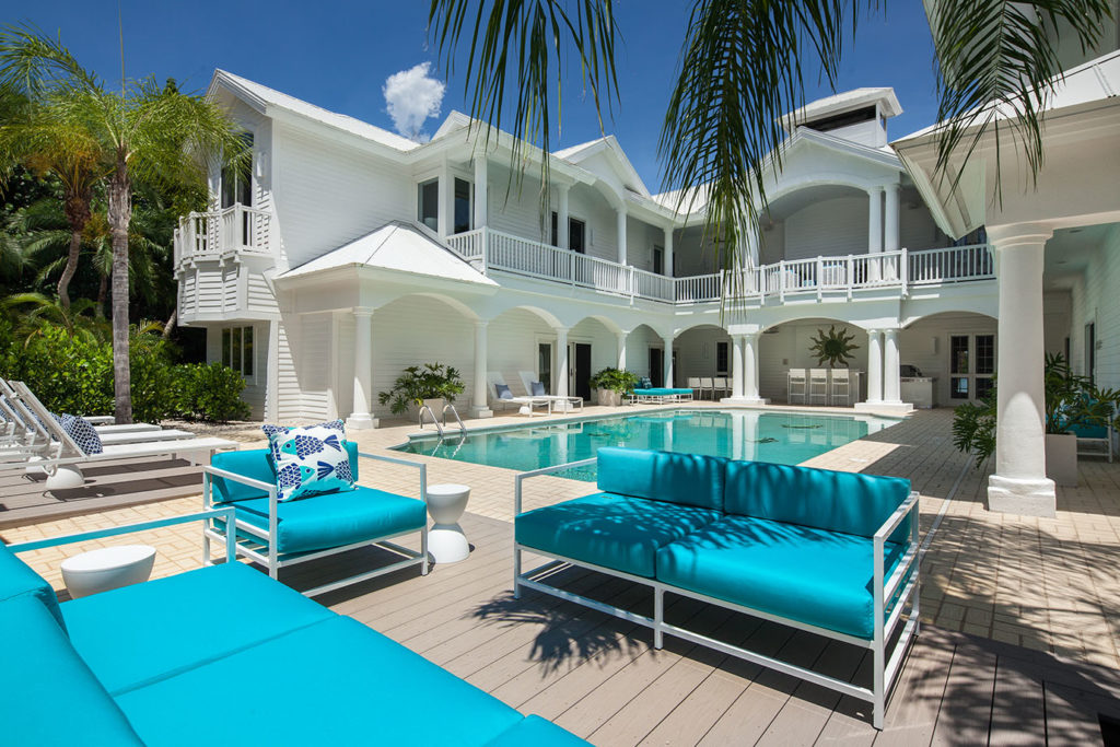 White Estate in Captiva Island, FL with an in-ground pool, blue patio furniture, white pool chairs, a patio area with green plants and a second floor balcony overlooking the pool surrounded by palm trees